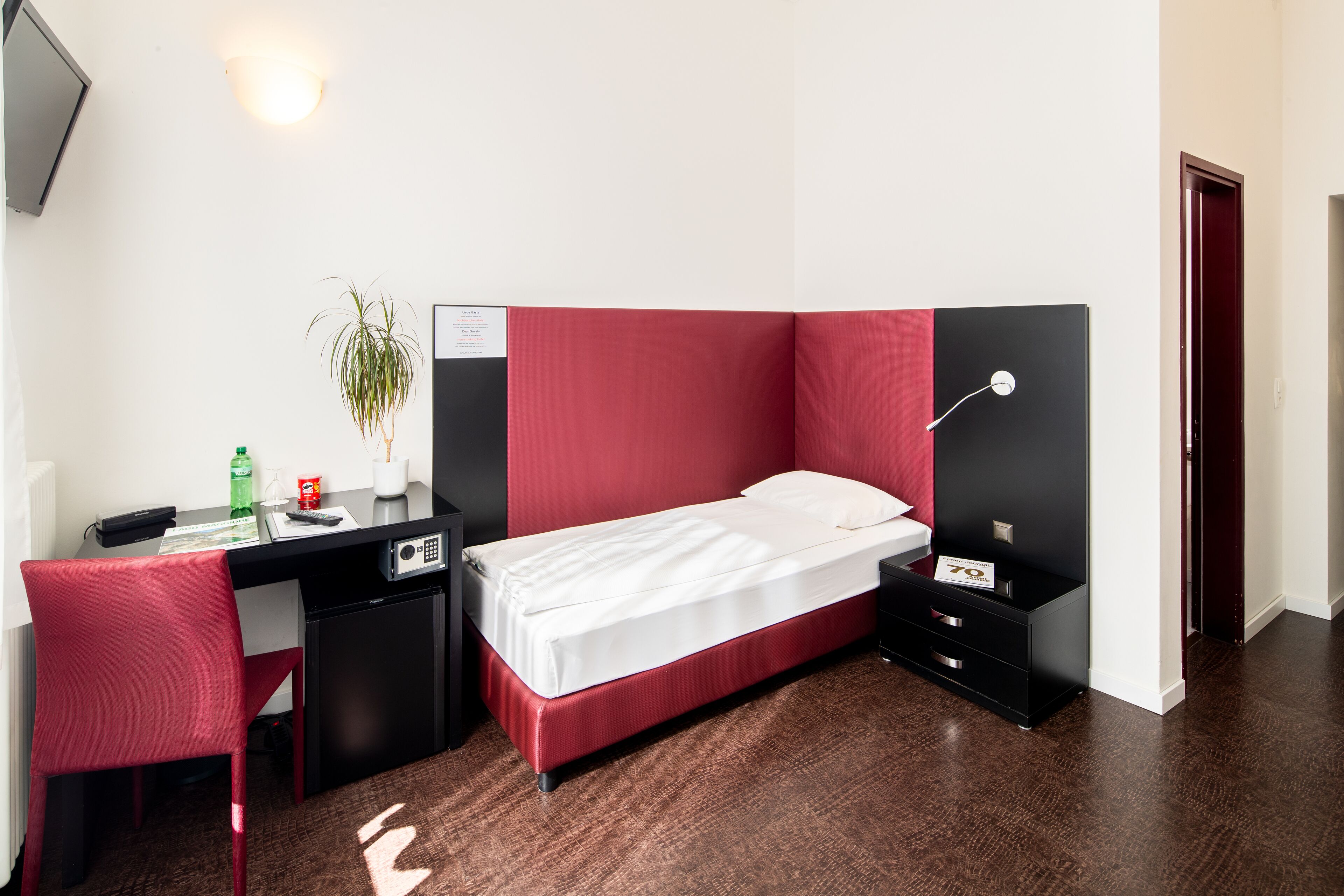 A compact hotel room with a single bed, a desk with a chair, a small desk lamp, and a nightstand with a lamp.
