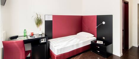 A compact hotel room with a single bed, a desk with a chair, a small desk lamp, and a nightstand with a lamp.