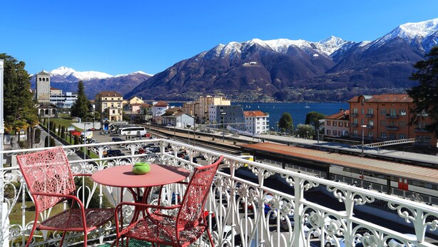 Un balcón con sillas metálicas rojas y una mesa, con vista a un pueblo con edificios y una cordillera.