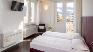 Habitación de hotel con cama, una silla, televisión y ventana con vistas a edificios y montañas.