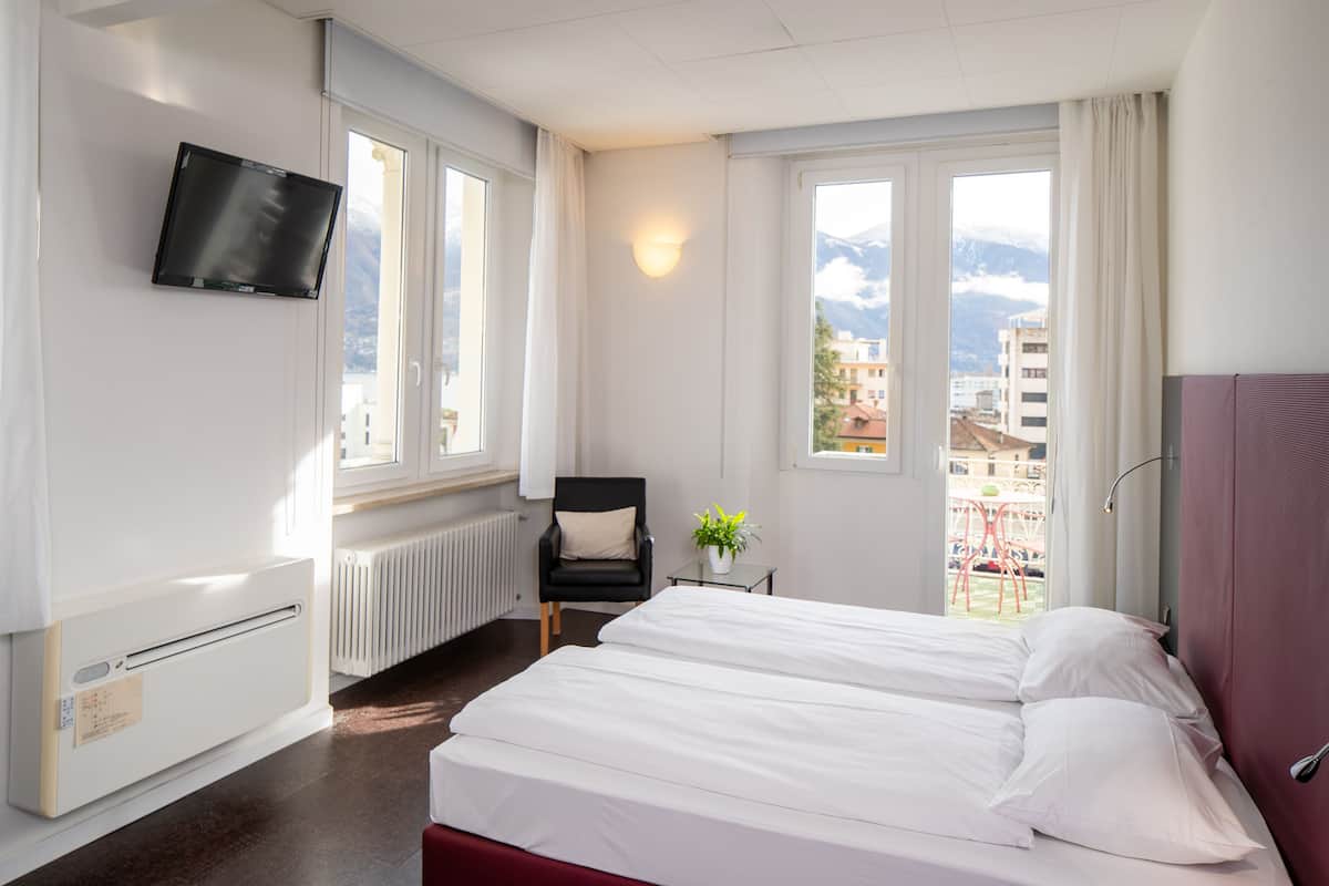 A hotel room with a bed, a chair, a TV, and a window with a view of buildings and mountains.