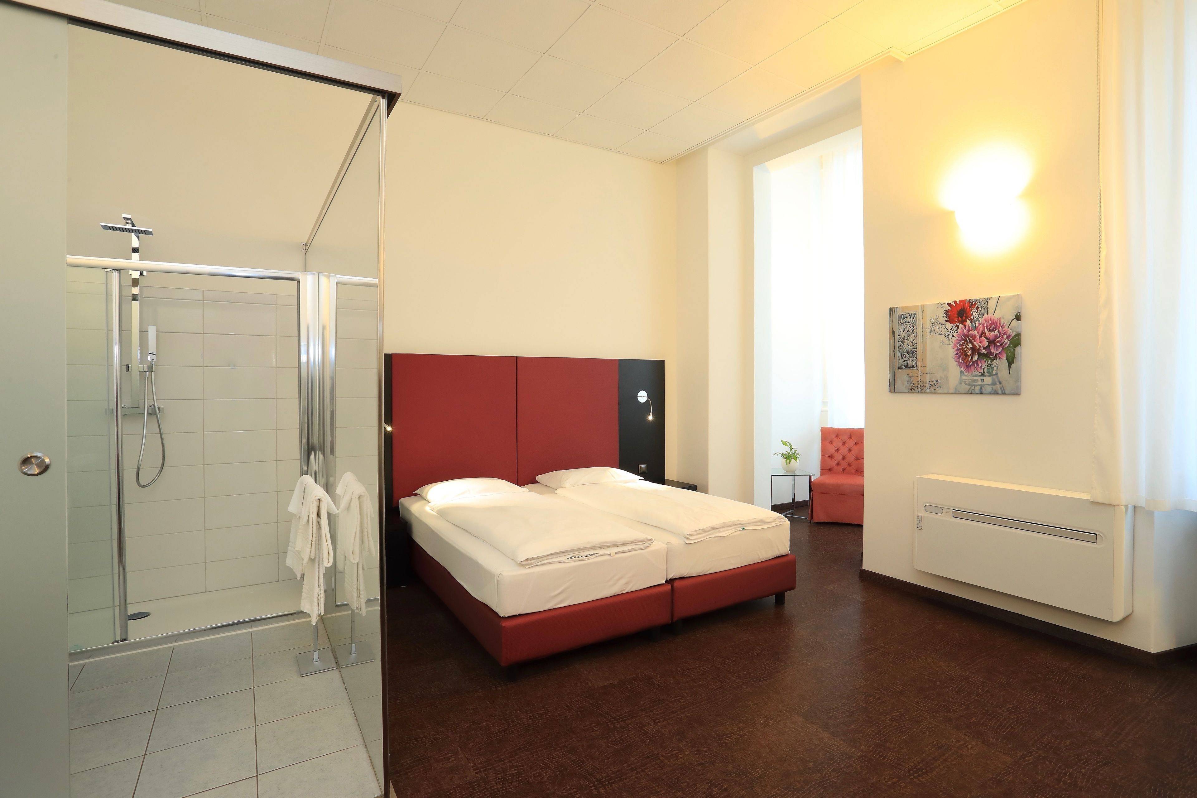 A modern hotel room with a glass shower enclosure, a bed with red headboard, and a white wall-mounted heater.