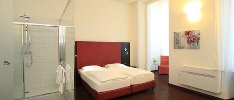 A modern hotel room with a glass shower enclosure, a bed with red headboard, and a white wall-mounted heater.