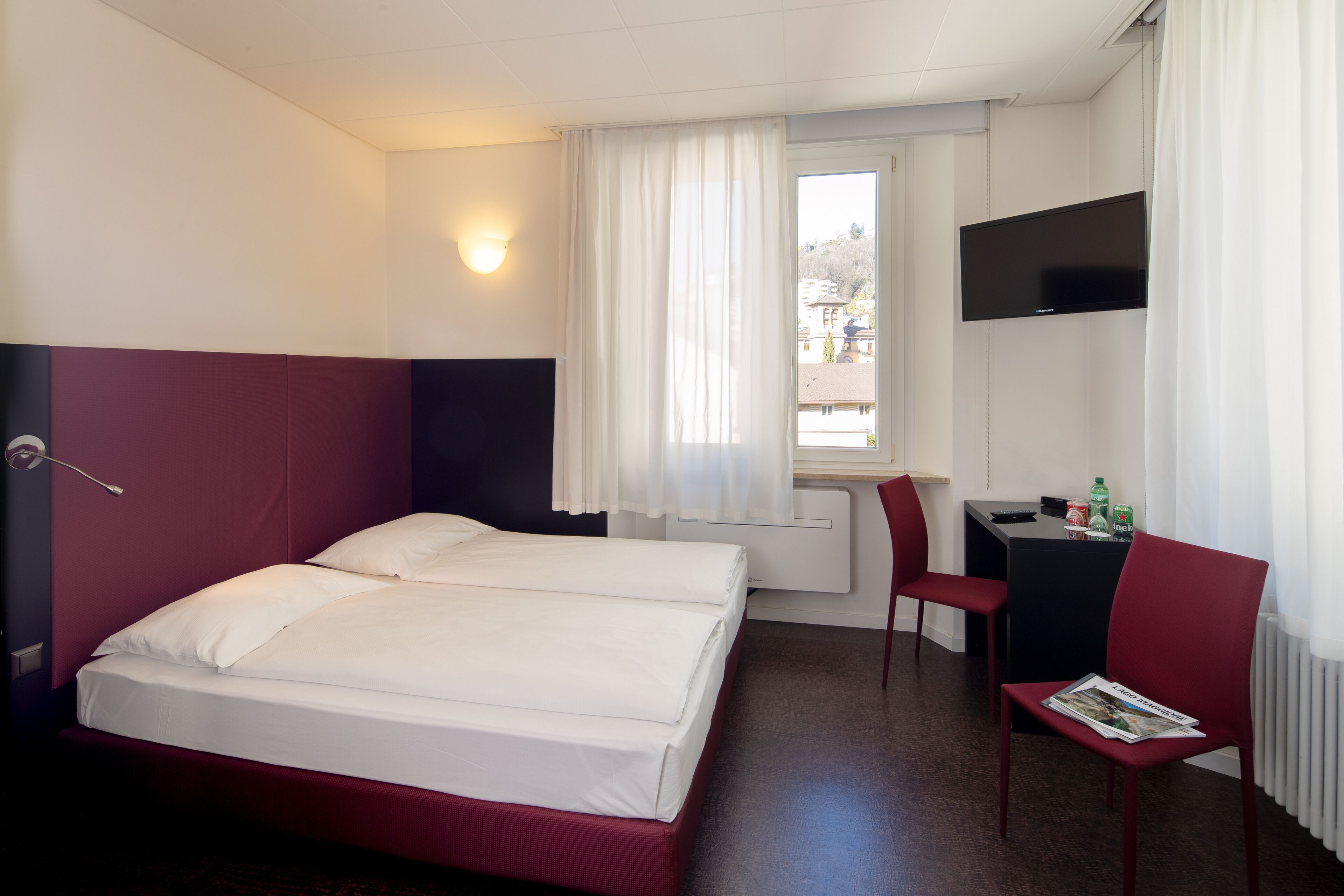 A hotel room with a bed, a desk, a chair, a TV, and a window with curtains.
