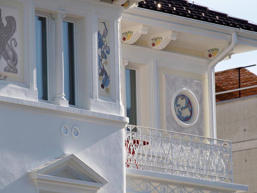 Un edificio con fachada blanca, molduras decorativas y un balcón con barandilla de metal ornamentada.