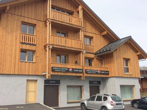 A multi-story wooden building with balconies, signs for rental and sales, and a parking area with cars. - 2-star apartment for 10 pers, 3 br, near ski lift, parking (La Toussuire)