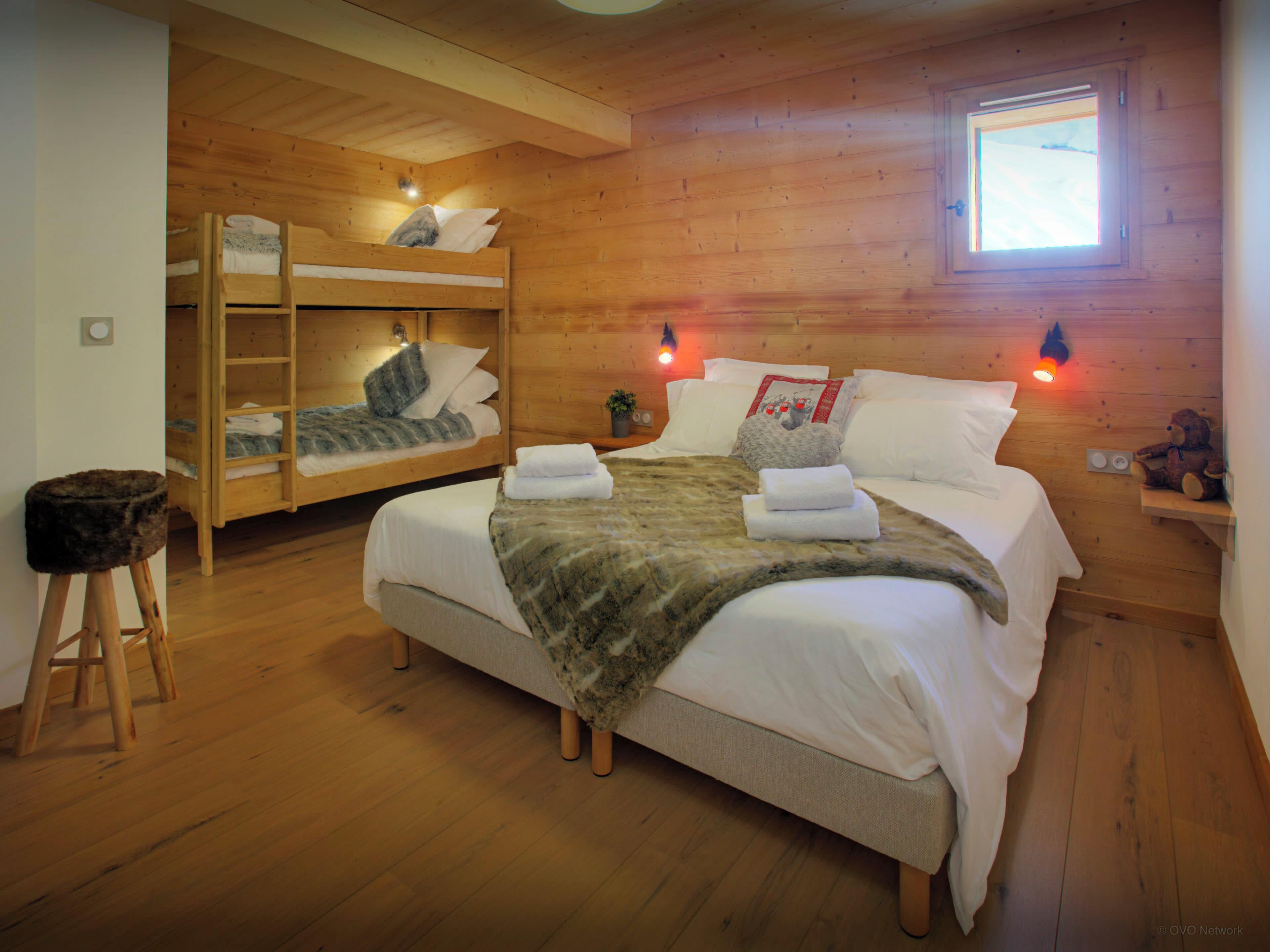 A wooden cabin bedroom with a large bed, two bunk beds, a small stool, and a window.