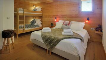 A wooden cabin bedroom with a large bed, two bunk beds, a small stool, and a window.