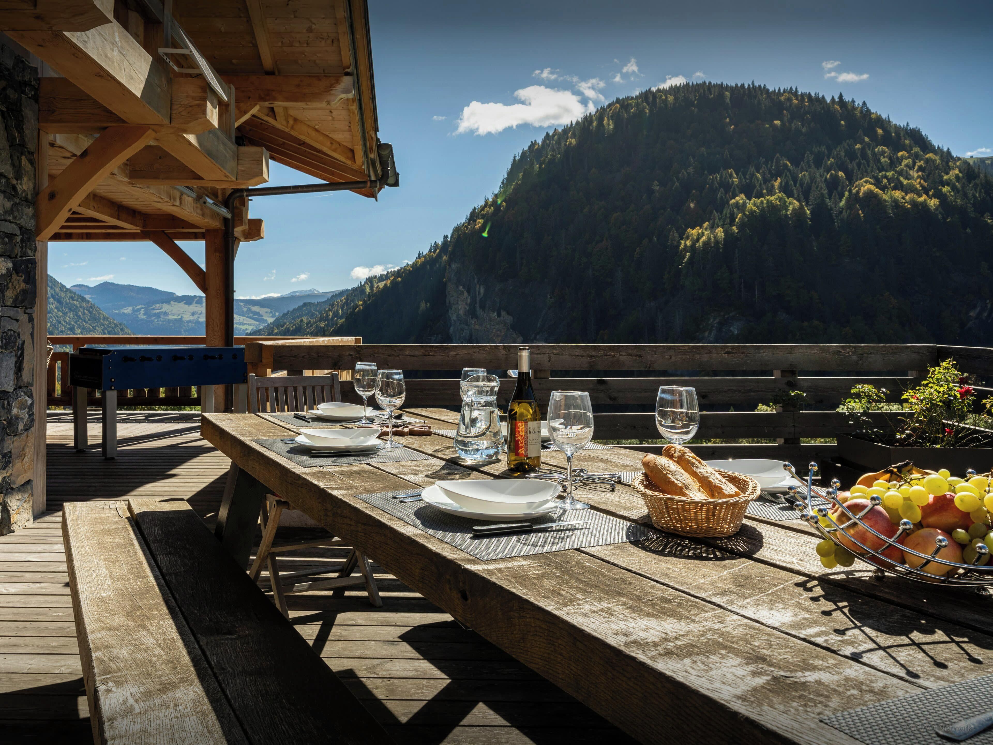 A wooden table set outdoors with a mountain view, glasses, a bottle, and a basket of fruit.