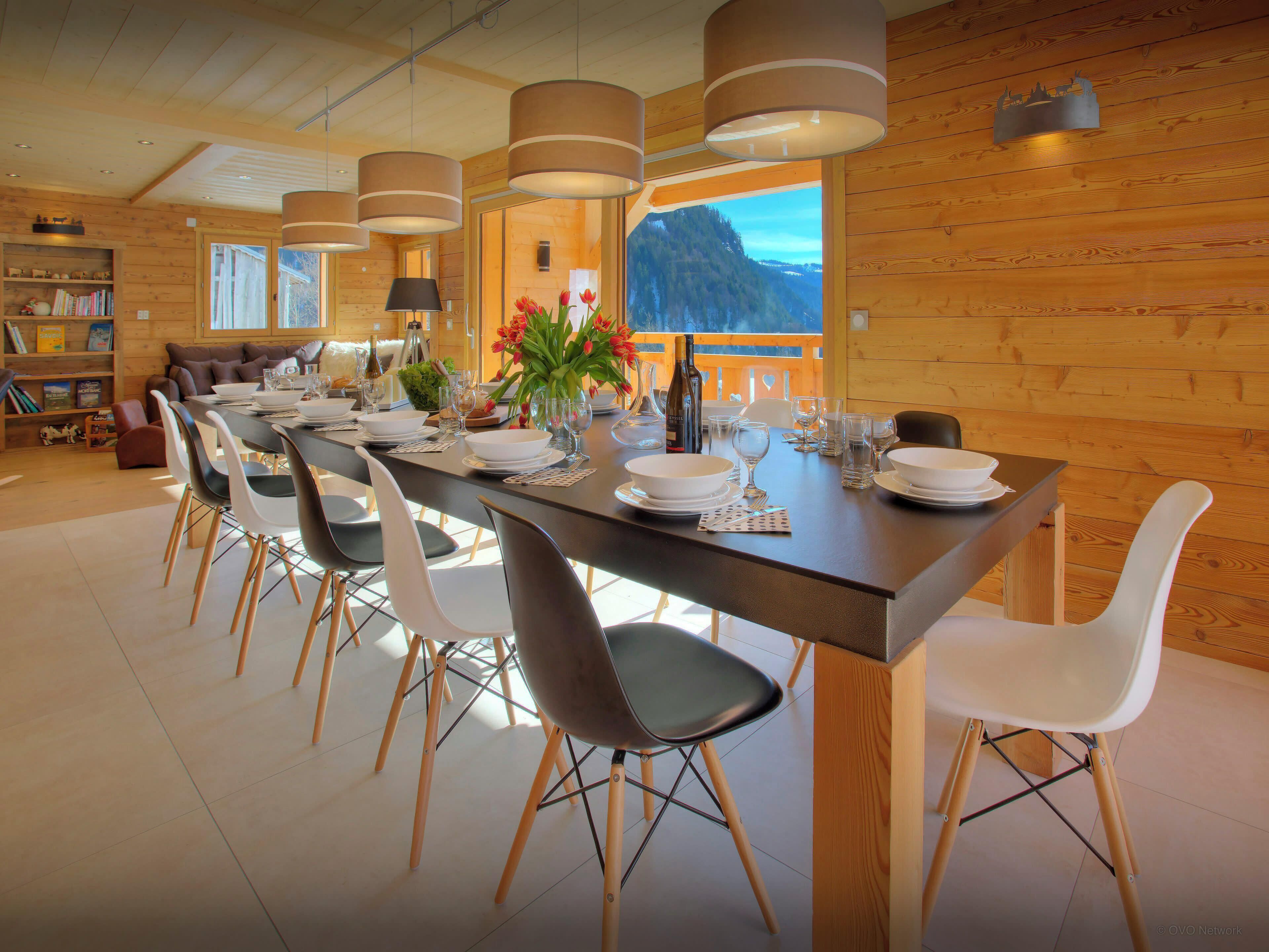 Uma sala de jantar com uma mesa comprida posta para seis pessoas, rodeada por cadeiras modernas, com vista para uma montanha através de grandes janelas.
