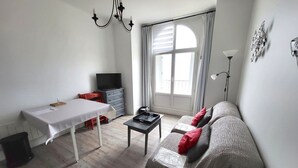 A living room with a sofa, a table, a TV on a dresser, and a large window with curtains. - Oceanfront 2-Room Apt with Direct Beach Access in Les Sables-d'Olonne (Les Sables-d'Olonne)