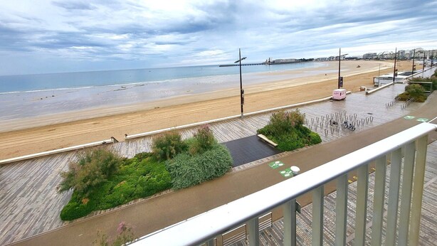 A beach view from a balcony with a wooden deck, a paved walkway, and a view of the sea and a bridge in the distance. - Oceanfront 2-Room Apt with Direct Beach Access in Les Sables-d'Olonne (Les Sables-d'Olonne)