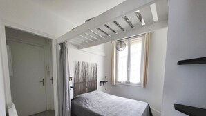 A bedroom with a bed, a window with curtains, a door, and a ceiling with a wooden beam structure. - Oceanfront 2-Room Apt with Direct Beach Access in Les Sables-d'Olonne (Les Sables-d'Olonne)