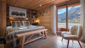 A wooden cabin bedroom with a bed, a bench, a fluffy chair, and a large window offering a mountain view. - Chalet Bonnevie - family home for 10, spa & mountain views - OVO Network (Les Villards-sur-Thônes)