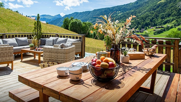 A wooden deck with outdoor furniture, a table with fruit and bowls, and a scenic mountain view. - Chalet Bonnevie - family home for 10, spa & mountain views - OVO Network (Les Villards-sur-Thônes)