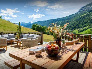 A wooden deck with outdoor furniture, a table with fruit and bowls, and a scenic mountain view.