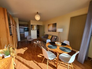 A modern living room with a dining area, wooden flooring, a ceiling light, a sofa with cushions, and a view of the kitchen in the background. - Studio near beach with private parking and balcony (Les Sables-d'Olonne)
