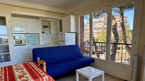 Ein kompakter Wohnraum mit Küchenzeile, einem blauen Sofa, einem kleinen Tisch und einem Balkon mit Blick auf Gebäude und Bäume.