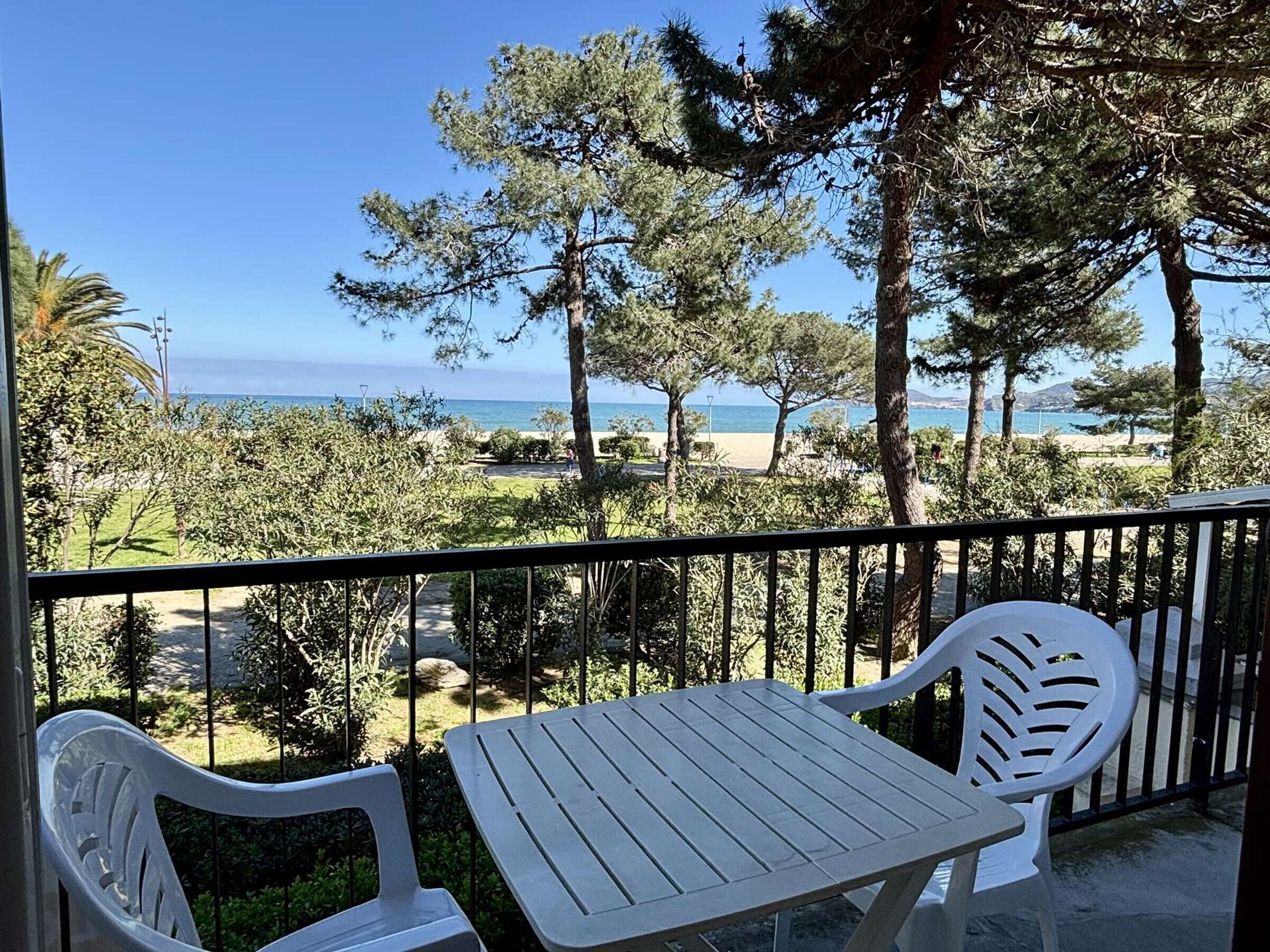 Un balcón con una mesa y dos sillas, con vista a una playa y un cielo despejado.