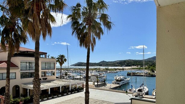 Un port de plaisance avec des bateaux amarrés, un bâtiment portant l’enseigne « PIZZA TOP », et une vue sur la mer et les collines en arrière-plan.