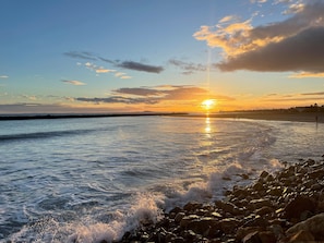 Escena de playa con el sol poniéndose en el horizonte, olas rompiendo sobre la orilla y una costa rocosa.