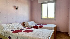 Une chambre avec deux lits en bois, chacun recouvert d’une courtepointe blanche ornée de motifs circulaires rouges. Les murs sont peints en rose et présentent un motif floral discret. Une fenêtre offre une vue sur la verdure à l’extérieur.