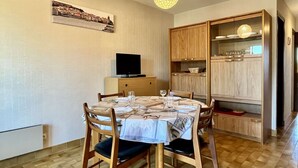Un coin repas avec une table ronde dressée pour quatre personnes, des chaises en bois, un téléviseur posé sur un meuble et un luminaire suspendu.