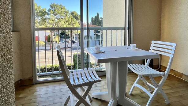 Un balcon avec une table et des chaises blanches, donnant sur un parking, et un bâtiment visible à l’arrière-plan.