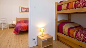 A dormitory room with bunk beds, a bedside table with a lamp, a chair, and a framed picture on the wall.