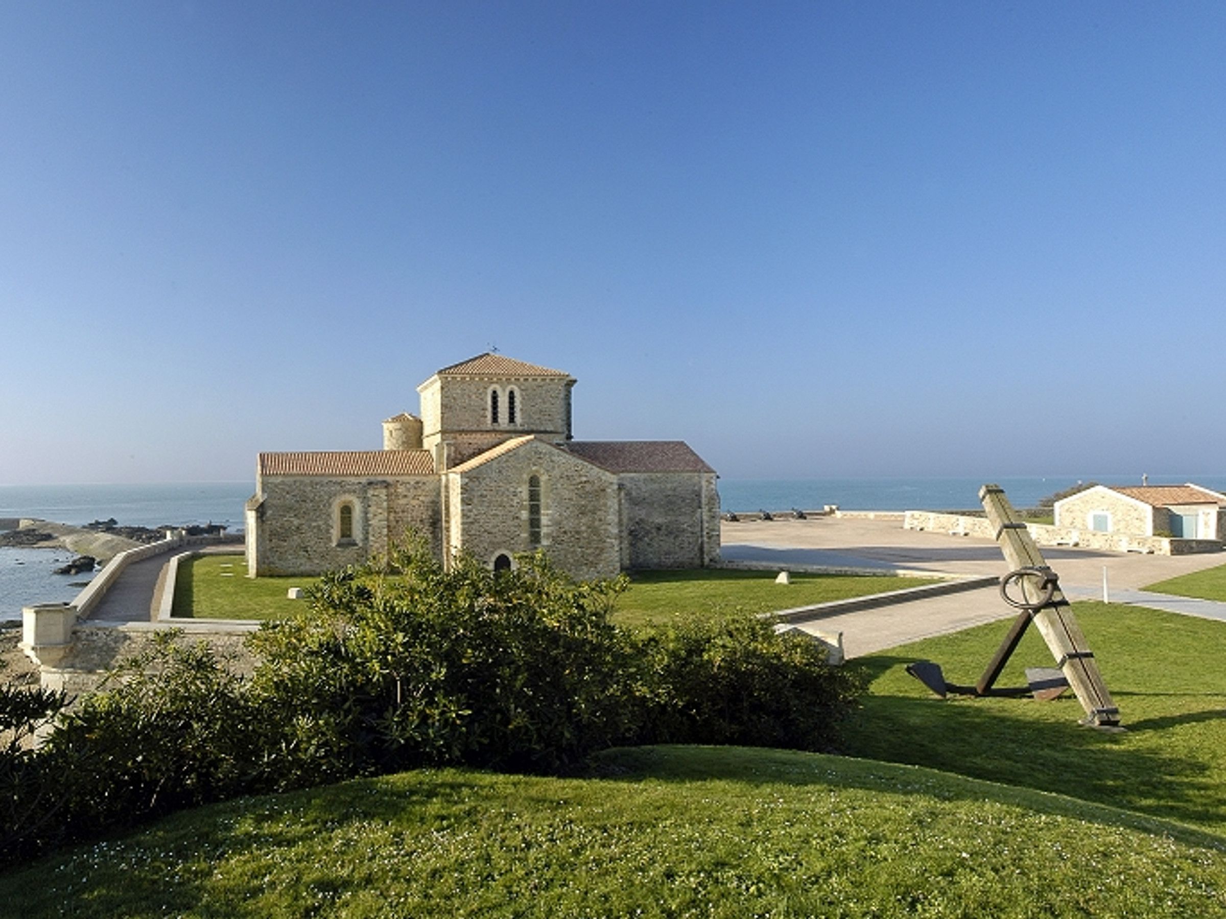 A stone church with a bell tower, located near a coastal area with a clear sky.