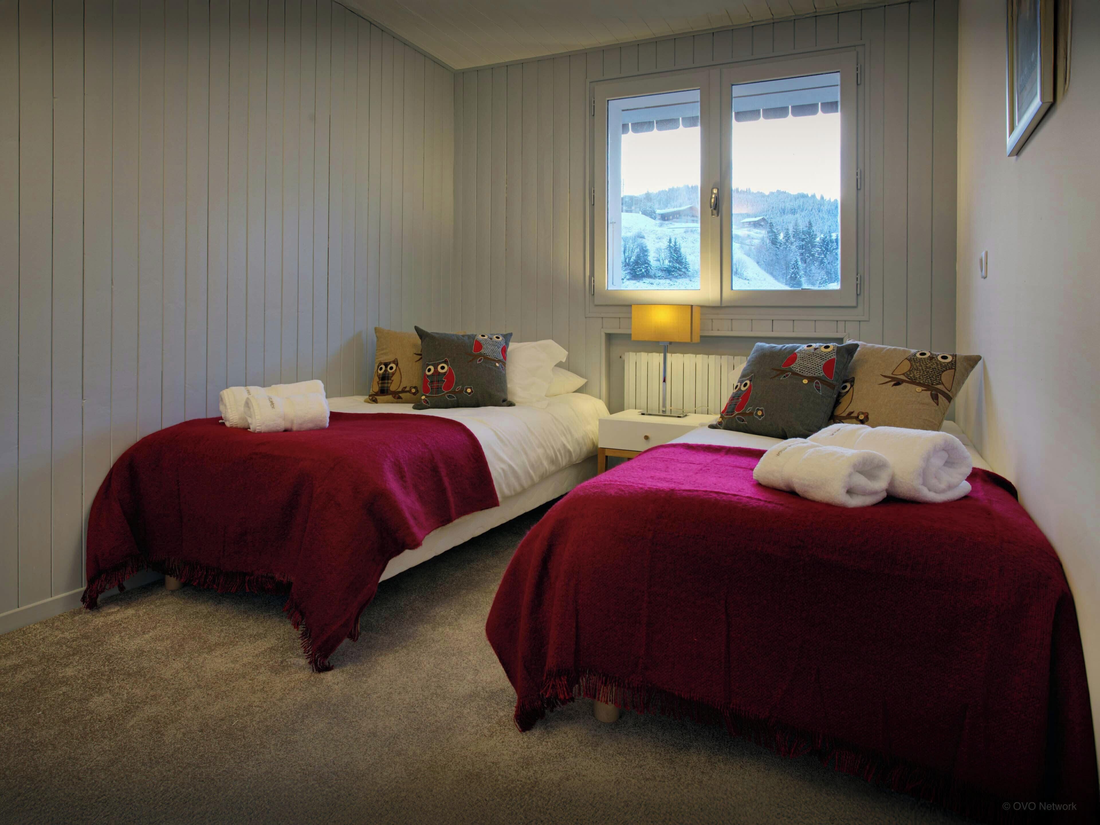 A hotel room with two beds, each with a red blanket and white towels, a nightstand with a lamp, and a window with a view of a snowy landscape.