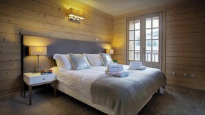 A neatly made bed with white linens and a decorative throw, a bedside table with a lamp, and a window with a view of a snowy landscape. - Chalet Le Mousqueton - ski base for 14 spa & mountain views - OVO Network (La Clusaz)