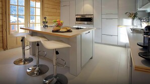 A modern kitchen with a central island, white cabinetry, stainless steel appliances, and a wooden door with windows. - Chalet Le Mousqueton - ski base for 14 spa & mountain views - OVO Network (La Clusaz)