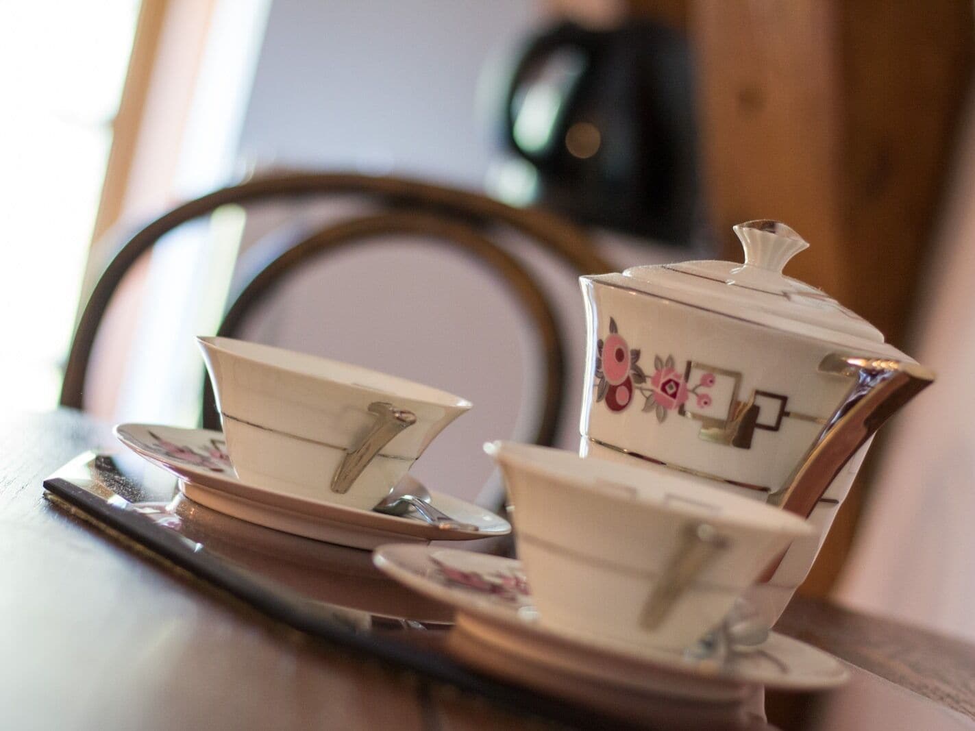 A tea set with a teapot and two cups on a saucer, placed on a table with a chair in the background.