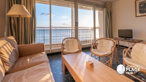 Een hotelkamer met een bank, twee rieten stoelen, een houten tafel, een televisie en een groot raam met uitzicht op zee.