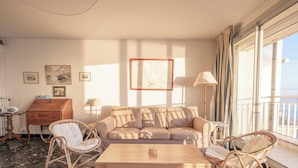 Een ruime woonkamer met een beige bank, rieten stoelen, een houten salontafel en een groot raam met uitzicht op zee.