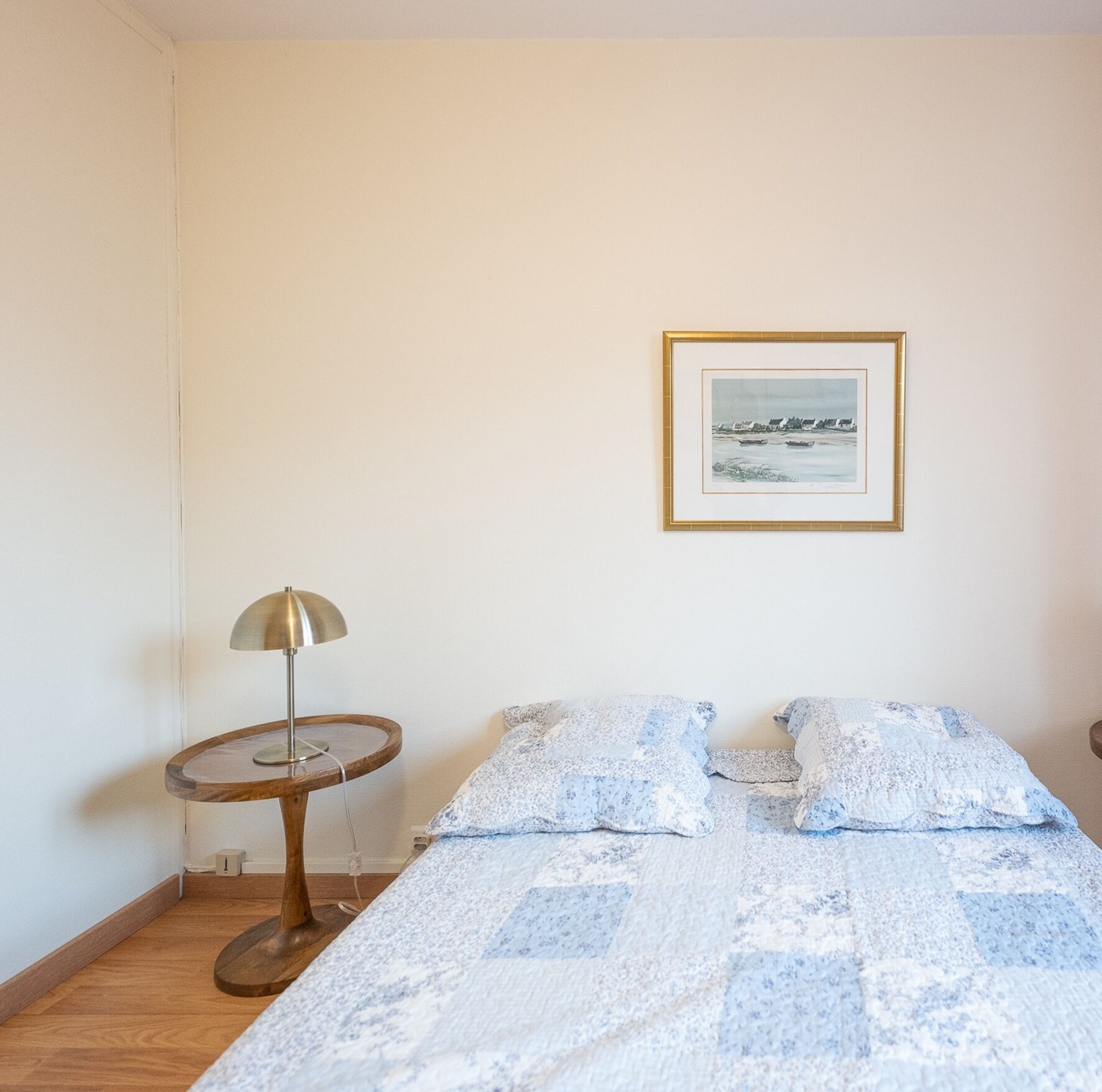 Un dormitorio con una cama, una mesa redonda con lámpara y un cuadro en la pared.