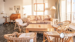 Een ruime woonkamer met een beige bank, rieten fauteuils, een houten salontafel en een piano.