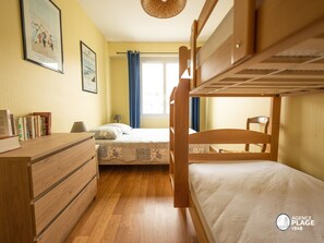 Una habitación con literas, un aparador de madera, una mesita de noche y una lámpara. Hay cuadros enmarcados en la pared y una ventana con cortinas.