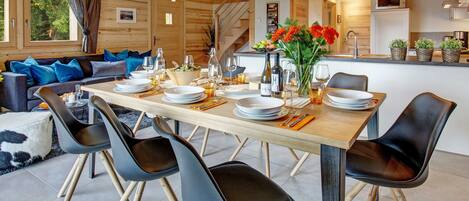 A dining area with a wooden table set for six, surrounded by black chairs. A wooden staircase is visible in the background, leading to an upper level.