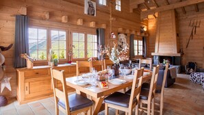 Interior de una cabaña de madera con comedor, paredes de madera, ventanas amplias, chimenea y una zona de estar acogedora.