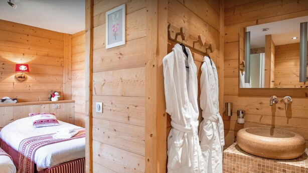 A wooden room with a bed, a bathroom with a round sink, and two white robes hanging on hooks.
