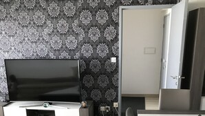 A modern living room with a flat-screen TV on a white entertainment unit, a dining table with chairs, and a patterned wallpaper. - Time Away From Home (Asse)