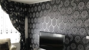 A modern living room with a flat-screen TV on a wooden stand, a dining table with chairs, a sofa, and patterned wallpaper. - Time Away From Home (Asse)