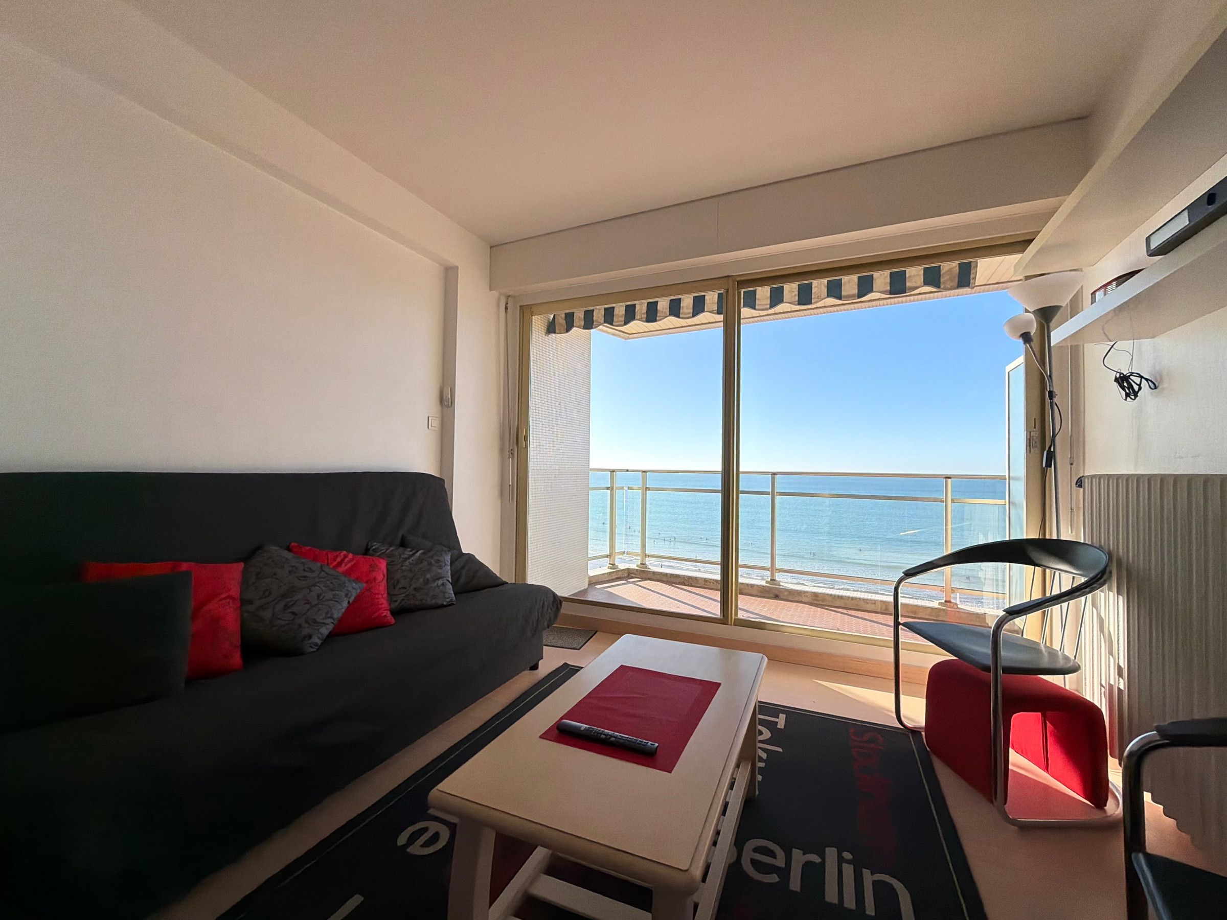 A room with a sofa, a small table, a chair, and a view of the sea through a sliding door.