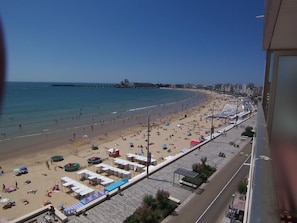 Uma praia com uma fileira de cabanas de praia e guarda-sóis, um calçadão, e vista clara do mar e edifícios ao longe.