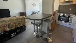 A compact kitchen with a built-in oven, a dining area with a round table and two bar stools, and a bed with a patterned cover.