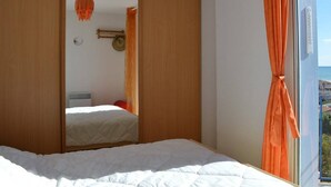 A bedroom with a large bed, a sliding wooden wardrobe, a mirror, and a view of the sea through a window with orange curtains. - 3-room villa with air conditioning, parking, and pets allowed in Saint-Pierre-la-Mer (Fleury-Saint-Pierre-la-Mer)