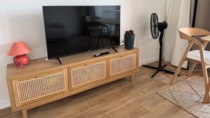 Un salon avec un téléviseur posé sur une console en bois, un ventilateur, un fauteuil et une lampe.