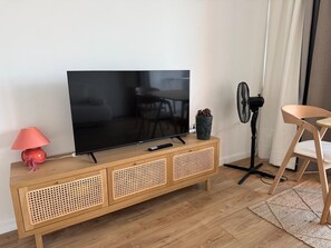 Uma sala de estar com televisão sobre um console de madeira, um ventilador, uma poltrona e um abajur.